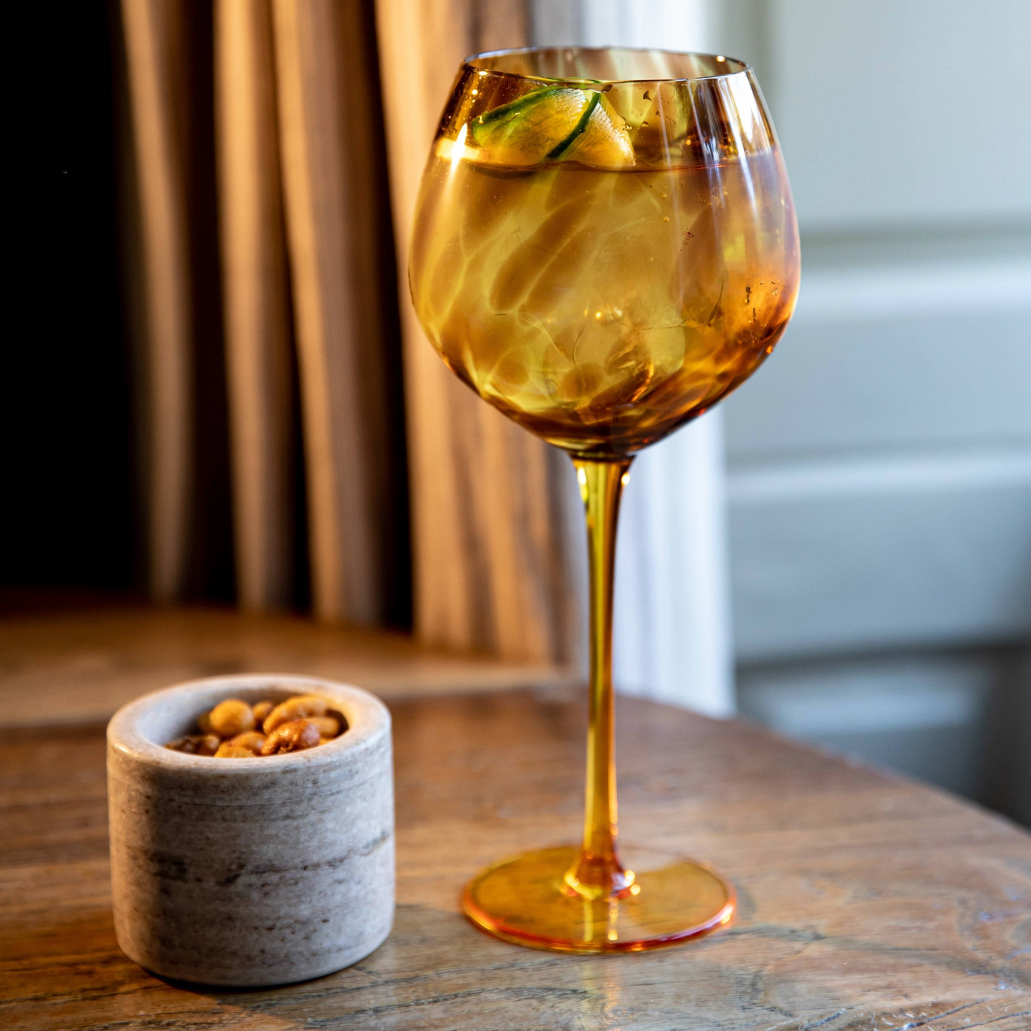 Sfeervolle setting met een gevuld gin glas op tafel, omringd door warme verlichting en een marmeren snackpot. Verkrijgbaar bij Sherlocks Place.