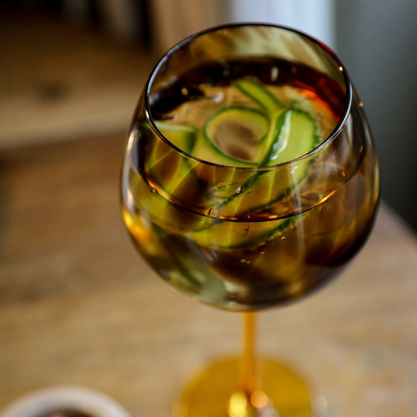 Bovenaanzicht van gin glas gevuld met een cocktail en verse komkommerlinten, tegen een houten tafel. Verkrijgbaar bij Sherlocks Place.