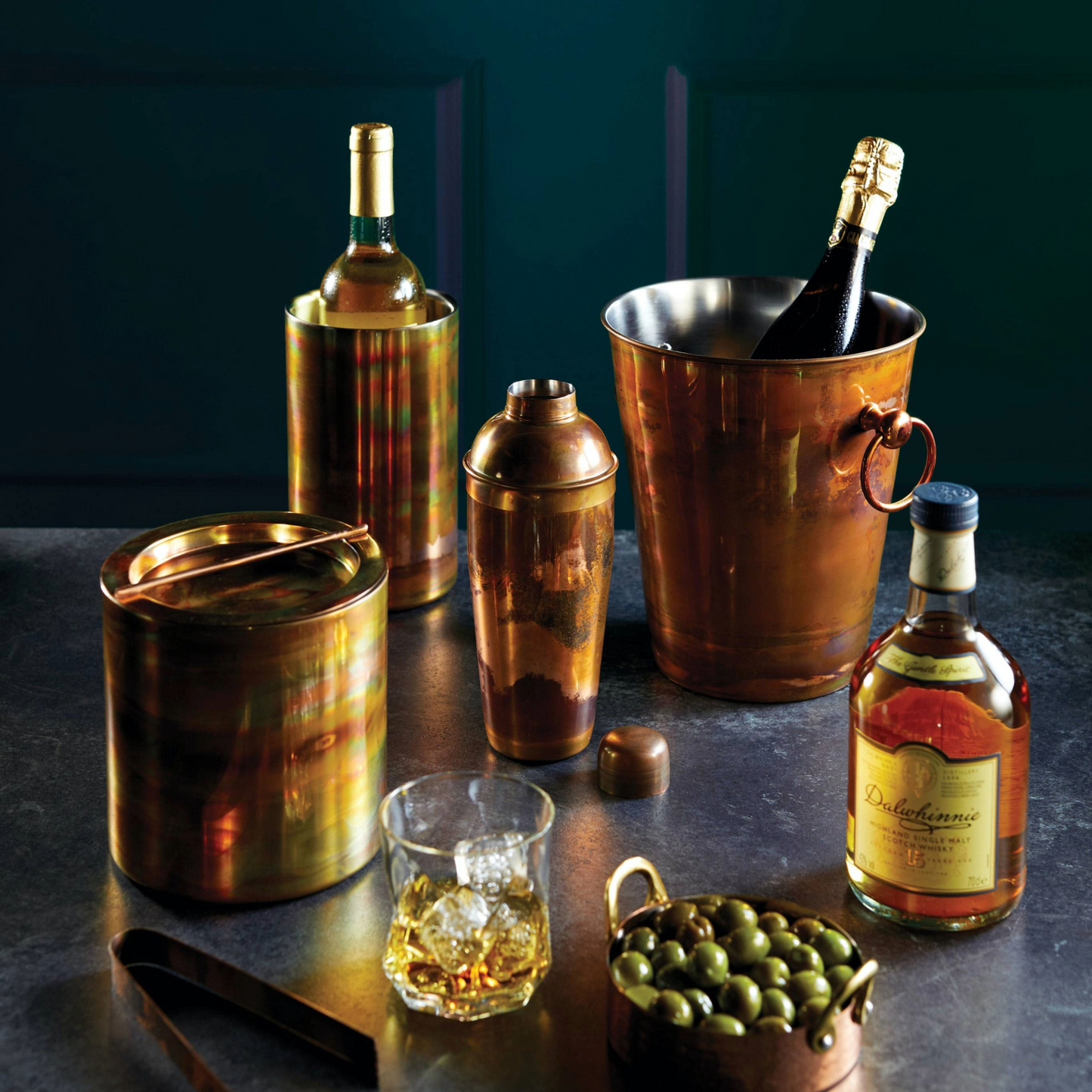 Sfeervolle barset met koperkleurige accessoires zoals wijnkoeler, ijsemmer en shaker met regenboogglans, op een donkere ondergrond – Verkrijgbaar bij Sherlocks Place
