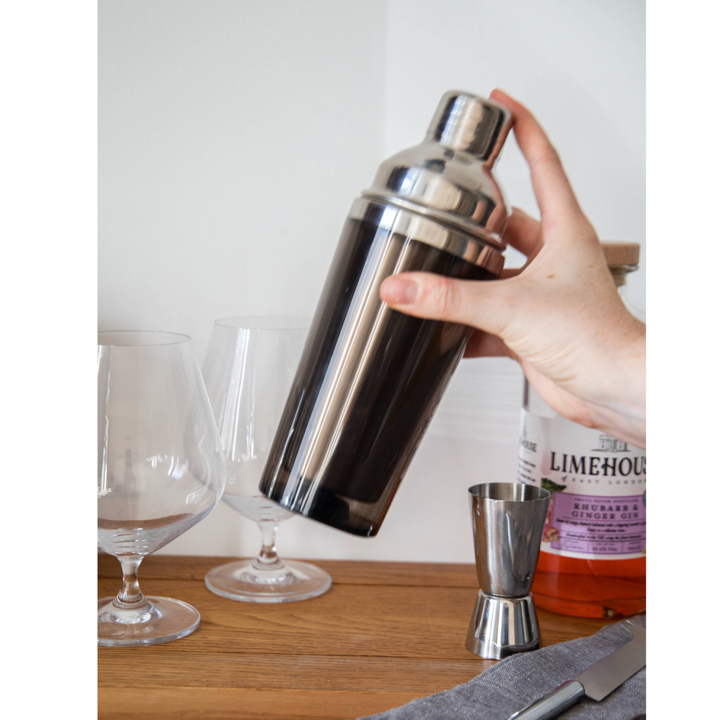BarCraft cocktailshaker stevig vastgehouden, met gin glazen en jigger op de achtergrond – essentieel accessoire voor mannen en bartenders die stijlvol willen shaken. Verkrijgbaar bij Sherlocks Place.