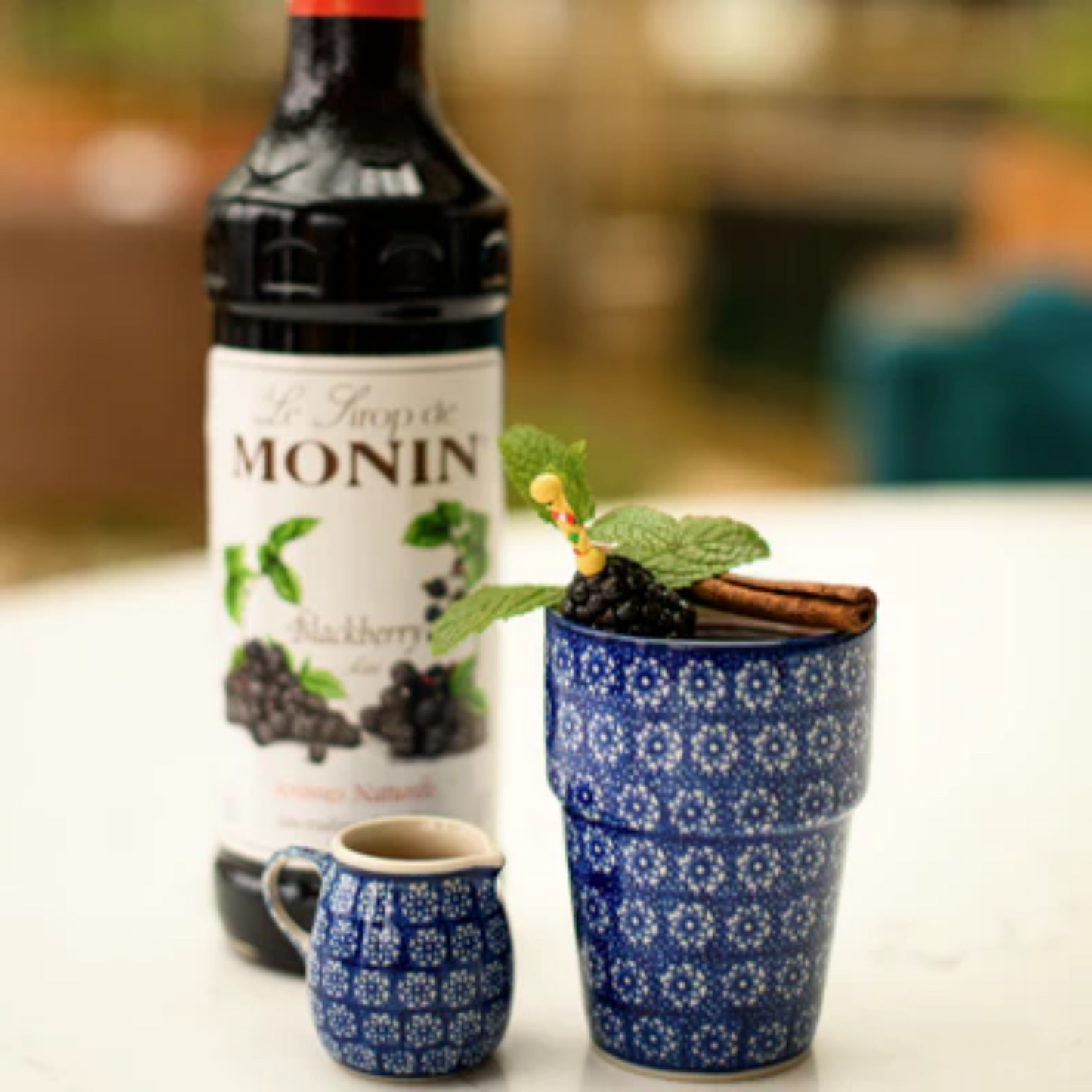 Een fles Monin Blackberry naast een blauwe keramische mok met glühwein, gegarneerd met muntblaadjes, bramen en een kaneelstokje, en een bijpassend klein kannetje. Monin verkrijgbaar bij Sherlocks Place Rotterdam