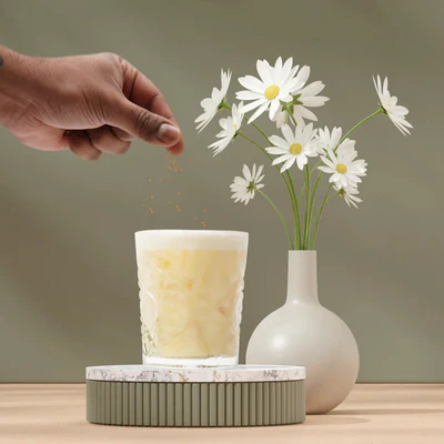 Een hand strooit nootmuskaat bovenop een romige Eggnog Chaï Vanille cocktail in een sierlijk glas, met op de achtergrond een witte vaas gevuld met frisse madeliefjes op een houten tafel. De sfeer is zacht en lenteachtig, perfect passend bij Pasen. Bourbon Vanilla Chai Eggnog Sherlocks Place Rotterdam
