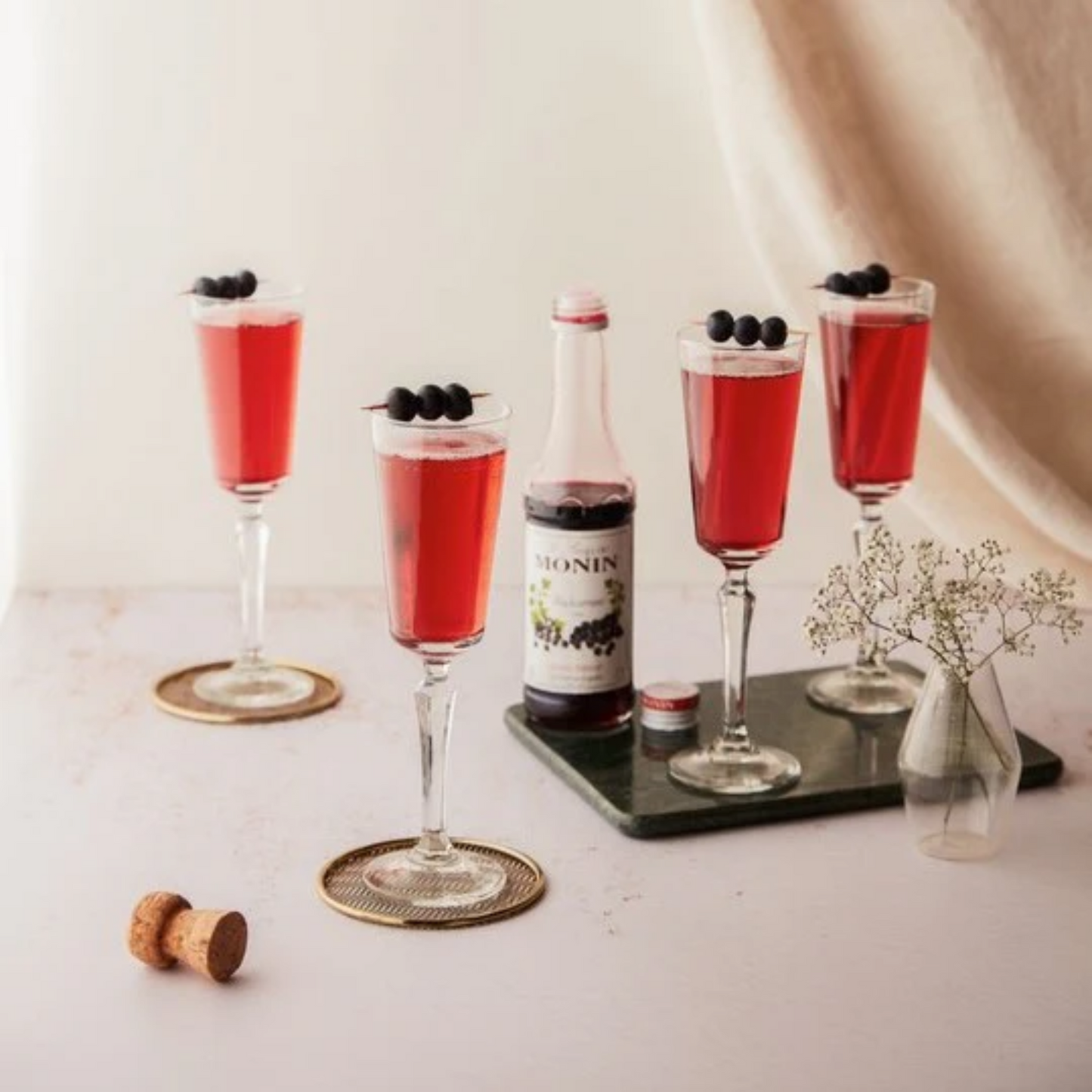 Een foto met 4 glazen Kir Royal gemaakt met Monin Cassis, ook wel Blackcurrant en Zwarte Bes genaamd. Teven een flesje Monin Cassis en een vaasje met bloemen is te zien. Als garnering op de glazen liggen cocktailprikkers met 3 olijven. Monin is verkrijgbaar bij Sherlocks Place Rotterdam