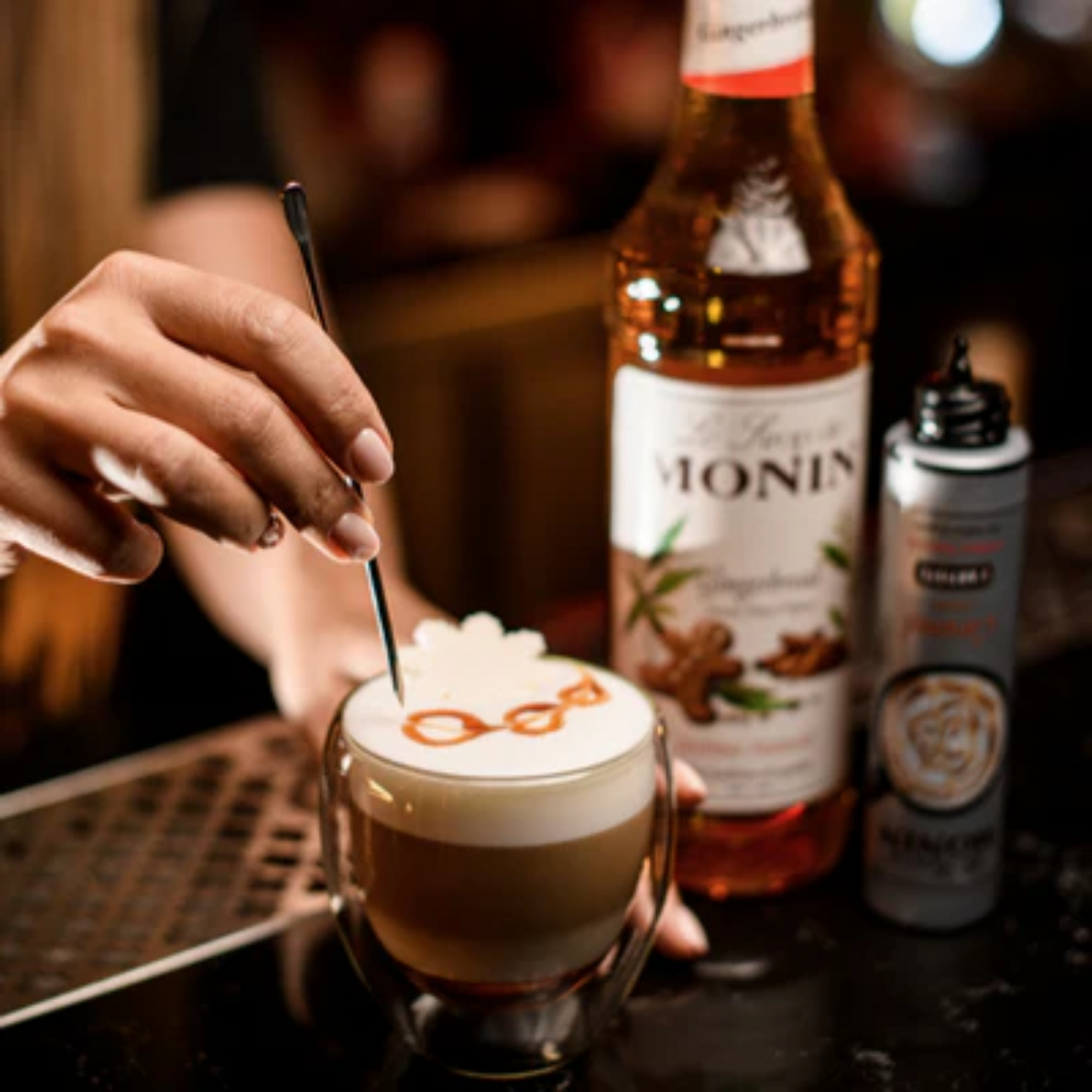 Barista maakt latte art met Monin Gingerbread siroop in een glas latte macchiato, met een fles Monin Gingerbread en toppingsiroop op de achtergrond. Verkrijgbaar bij Sherlocks Place Rotterdam.