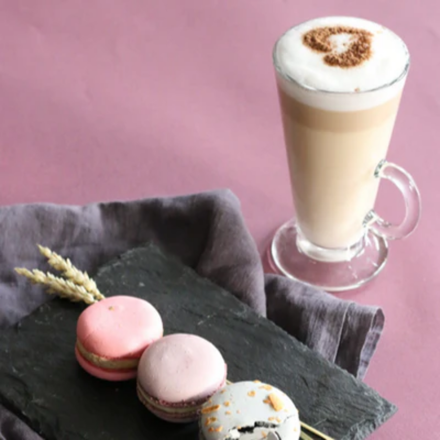 Romige latte met hartje van cacaopoeder, geserveerd met kleurrijke macarons waaronder een blackberry macaron op een leisteenbord, gecreëerd met MONIN siroop. Monin verkrijgbaar bij Sherlocks Place Rotterdam