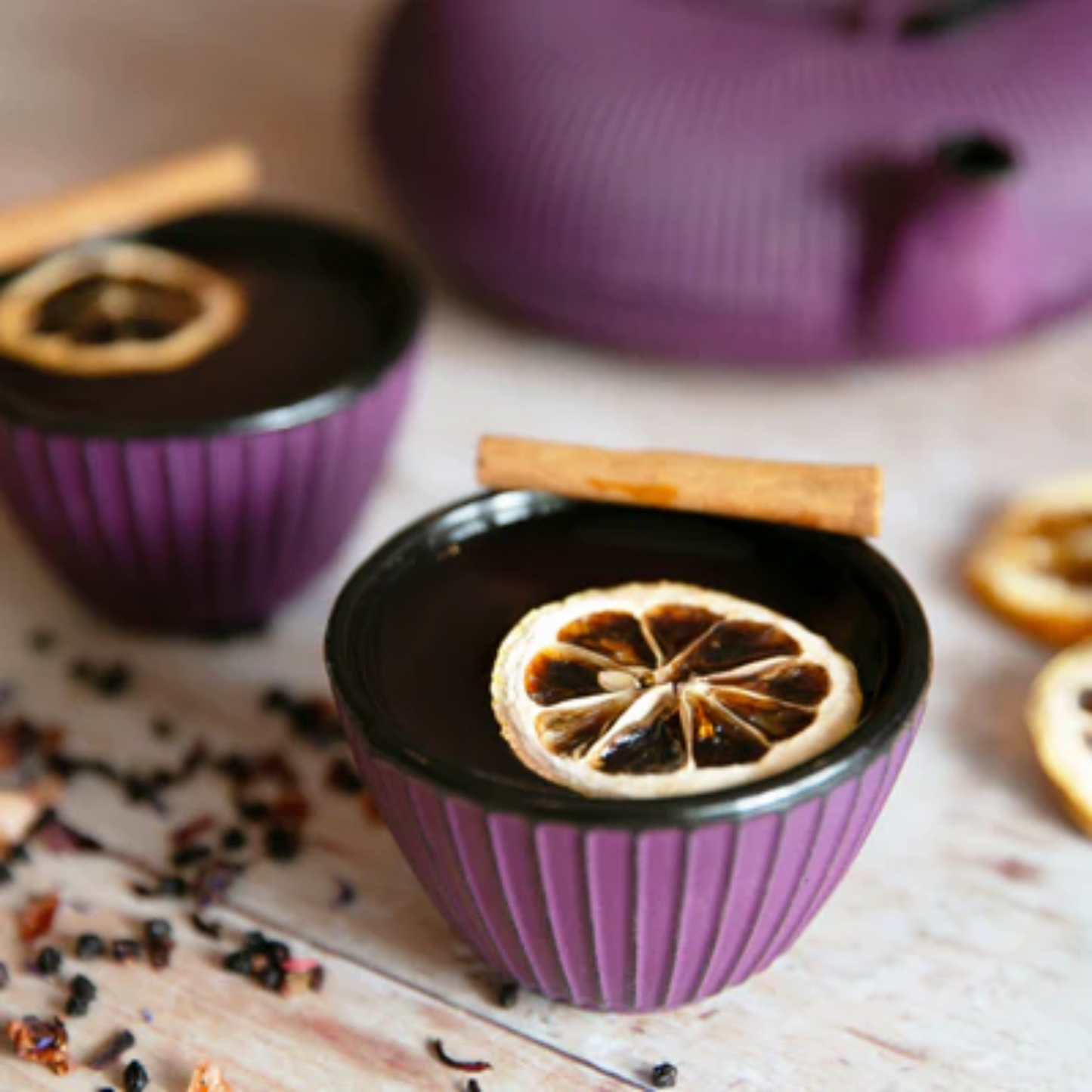 Mooie gedetailleerde Foto van twee Chinese theekopjes in de kleur paars. 1 kopje is uitgelicht en de andere en een paarse Chinese theepot zijn vager op de achtergrond. In de kopjes zit Chai Tea gemaakt met Monin Chai Tea concentraat 70cl. Verkrijgbaar bij Sherlocks Place Rotterdam. de thee is gegarneerd met een schijfje gedroogde citroen en een kaneelstokje