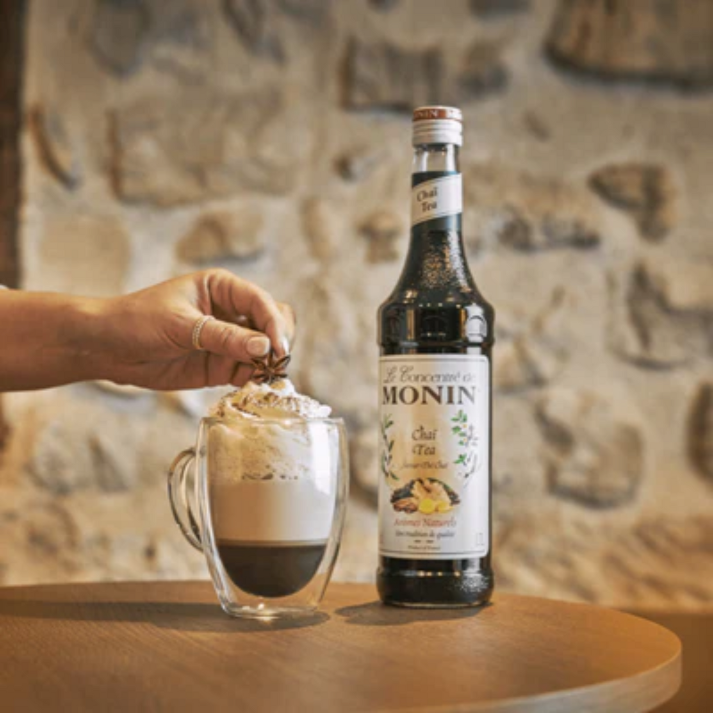 Mooi gedetailleerde foto van een glas met de unieke drank Coco Chai gemaakt van Monin producten. Het lijkt op een latte, onderin het glas de donkere kleur van de chai tea, daarboven een romige laag melk en daarboven een dot slagroom. Gegarneerd met wat kaneelpoeder en een steranijs. Het glas staat op een tafel met ernaast een fles Monin Chai Tea concentraat. Je ziet ook nog een hand het steranijs op de drank leggen. Monin verkrijgbaar bij Sherlocks Place Rotterdam