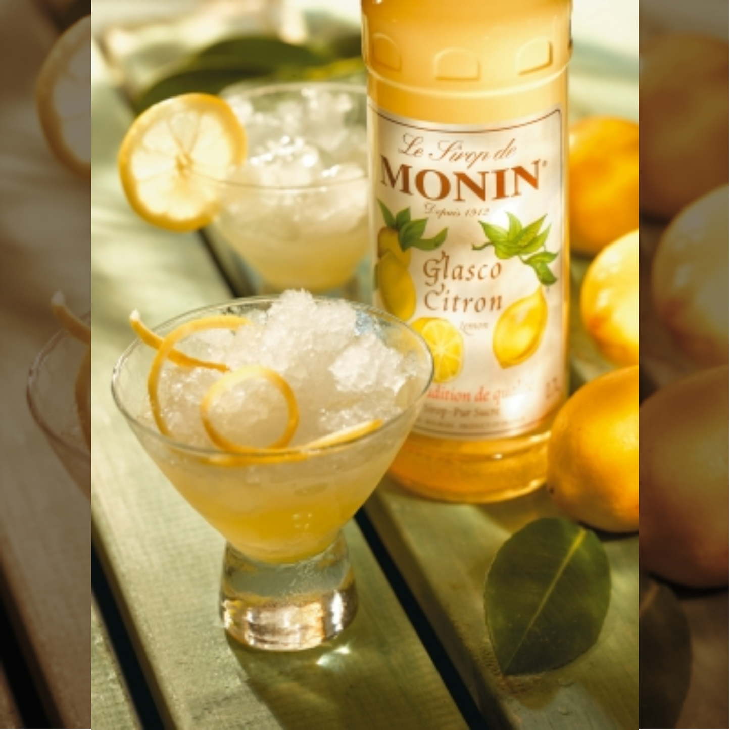 Een zonnige setting met twee glazen homemade lemonades naast een fles Monin Lemon Glasco. Het tafereel staat op een houten tuintafel en is verder gedecoreerd met citroenen. De galzen zijn gegarneerd met citrus schillen. Monin Verkrijgbaar bij Sherlocks Place Rotterdam. Must-have voor zon aanbidders en levensgenieters!