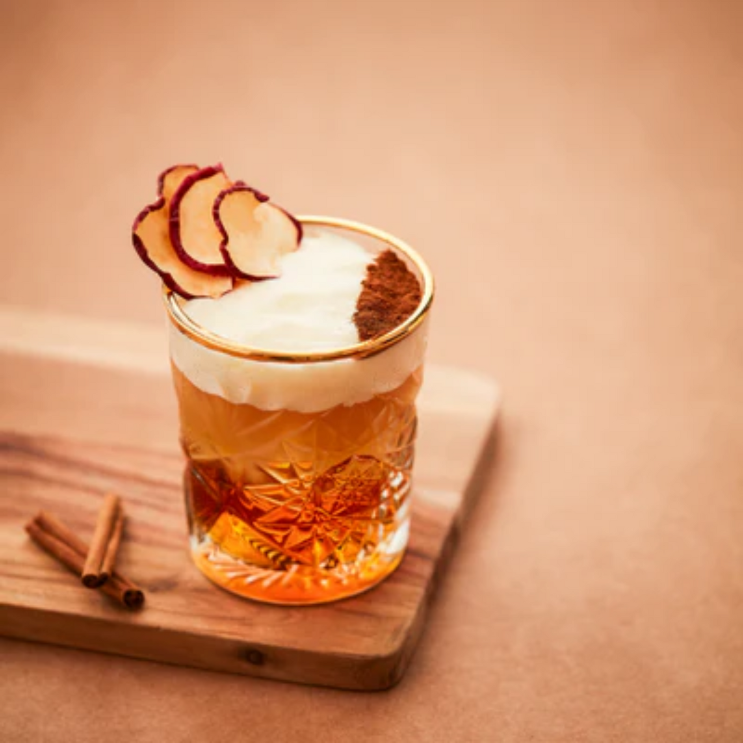Een glas Old Fashioned cocktail met een twist, gemaakt met onder andere Monin Speculoos siroop. De foto is in vintage look in bruintinten en het glas staat op een houten snijplank. De cocktail is gegarneerd met schijfjes gedroogde appel en kaneelpoeder. Op de plank liggen wat kaneelstokjes. Monin is verkrijgbaar bij Sherlocks Place Rotterdam