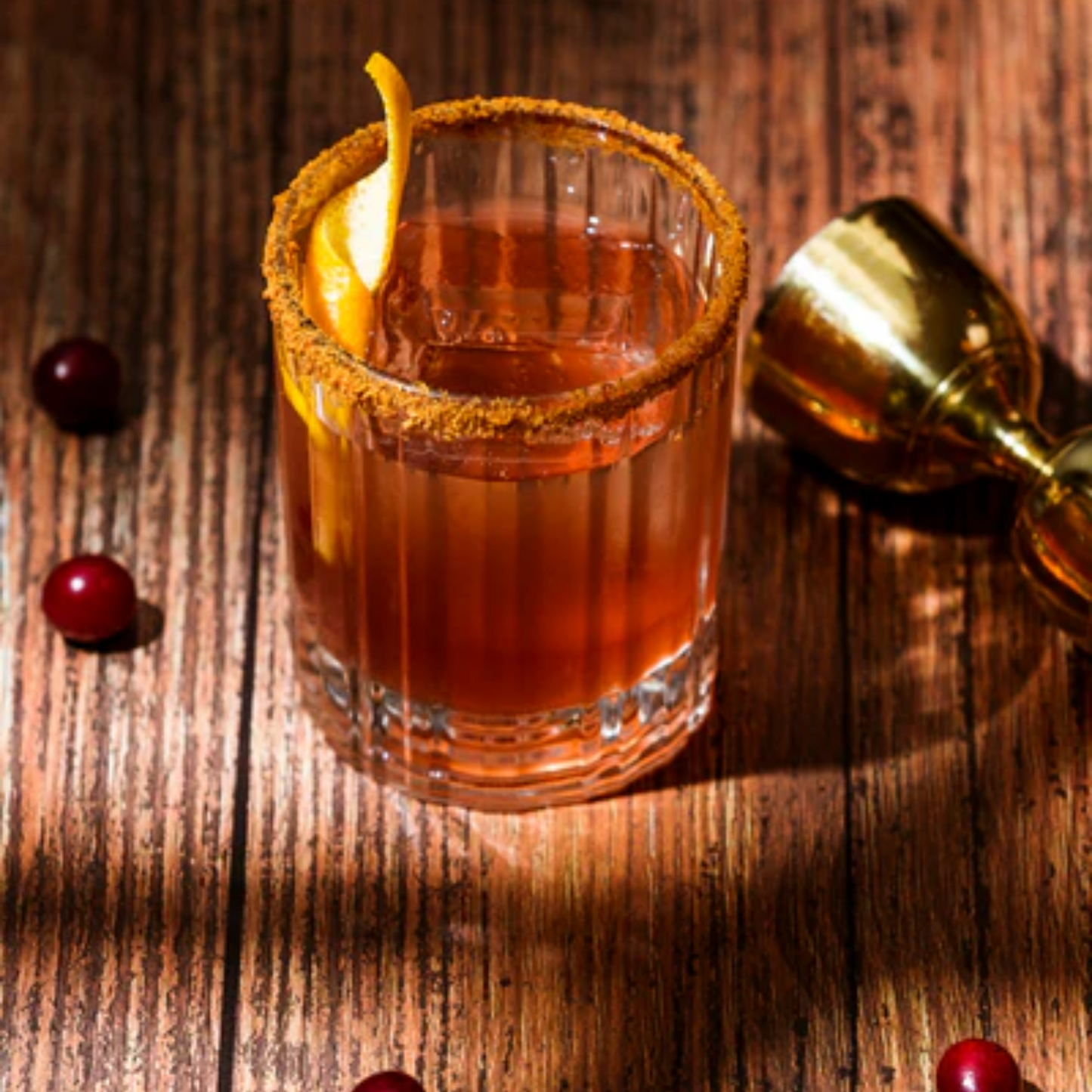 Een zeer mooie close-up foto van een Old Fashioned cocktail in vintage retro stijl. Een doorzichtig whiskey glas op een houten tafel met een bronskleurige jigger rechts en wat verse cranberries op de tafel. De Old Fashioned is gemaakt met Monin ingrediënten als Cranberry. Compleet Monin assortiment verkrijgbaar bij Sherlocks Place Rotterdam