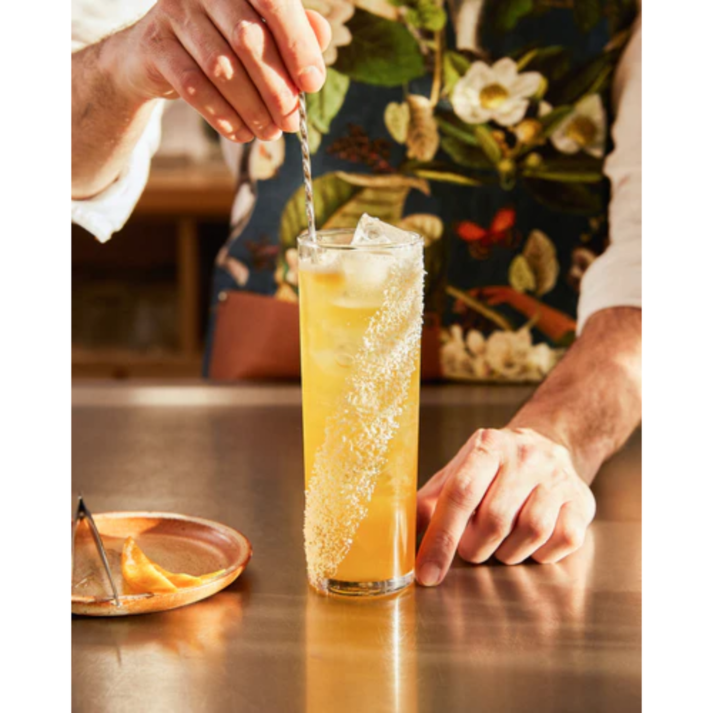 Close-up foto van een White Tea Mango Cocktail gemaakt met Monin. Het is een chique foto met op de achtergrond de bartender met een fleurig schort die de cocktail aan het bereiden is. Hij heeft een bar spoon in de hand. Op de voorgrond het glas; een longdrink gevuld met ijsklontjes en de Monin ingrediënten. De kleur is oranje. Ernaast een schaaltje met daarop een pincet en slices citrus. Alles van Monin verkrijgbaar bij Sherlocks Place Rotterdam