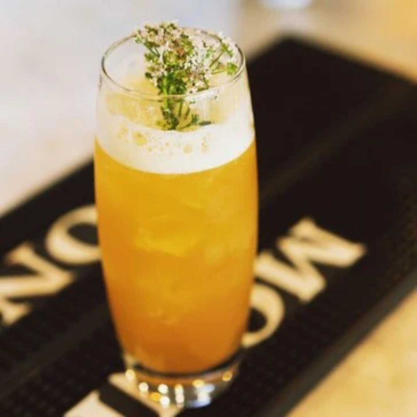 Bier in een hoog bierglas op een Monin barmat. Het bier heeft een mooie schuimkraag en in het schuim zijn wat takjes Vlierbloesem gestoken. Bier met bloemen? Jazeker! Met de Monin Elderflower siroop breng je elk biertje naar een next level! Met of zonder alcohol! Compleet Monin assortiment verkrijgbaar bij Sherlocks Place Rotterdam