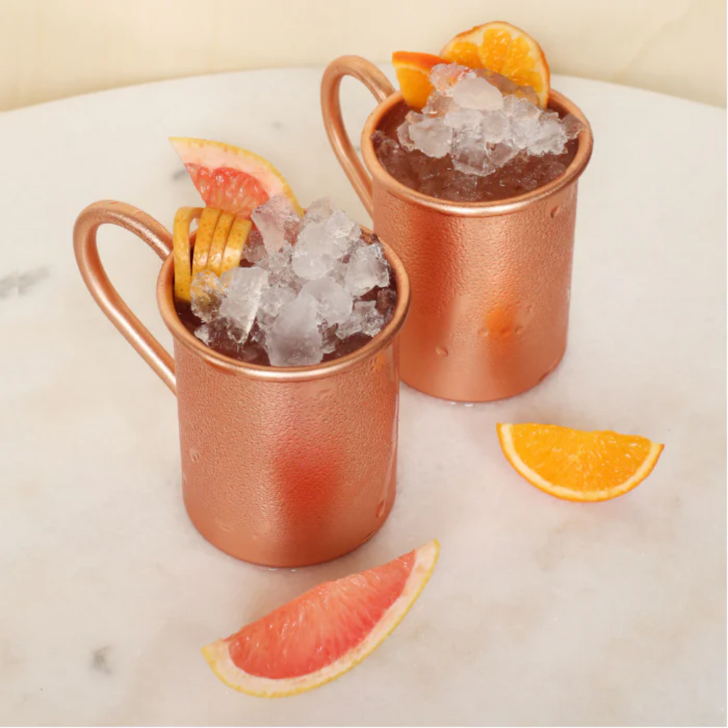 Twee unieke Moscow Mule bekers in de kleur koper op een witte tafel. Ervor liggen een partje rode grapefruit en een partje sinaasappel. De mules zijn gevuld met crushed ice en een moscow mule gemaakt van Monin en als garnering weer citrusvruchten. Monin verkrijgbaar bij Sherlocks Place Rotterdam