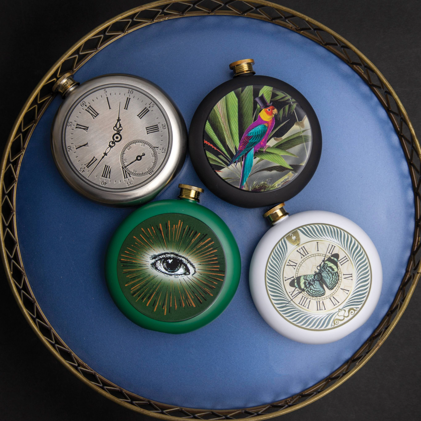 Vier ronde heupflessen met unieke designs, waaronder een zilverkleurige klok, een papegaai met hoed in een tropische setting, een groen oog met gouden stralen en een vlinder op een wijzerplaat, gepresenteerd op een blauwe ondergrond in een decoratieve schaal.