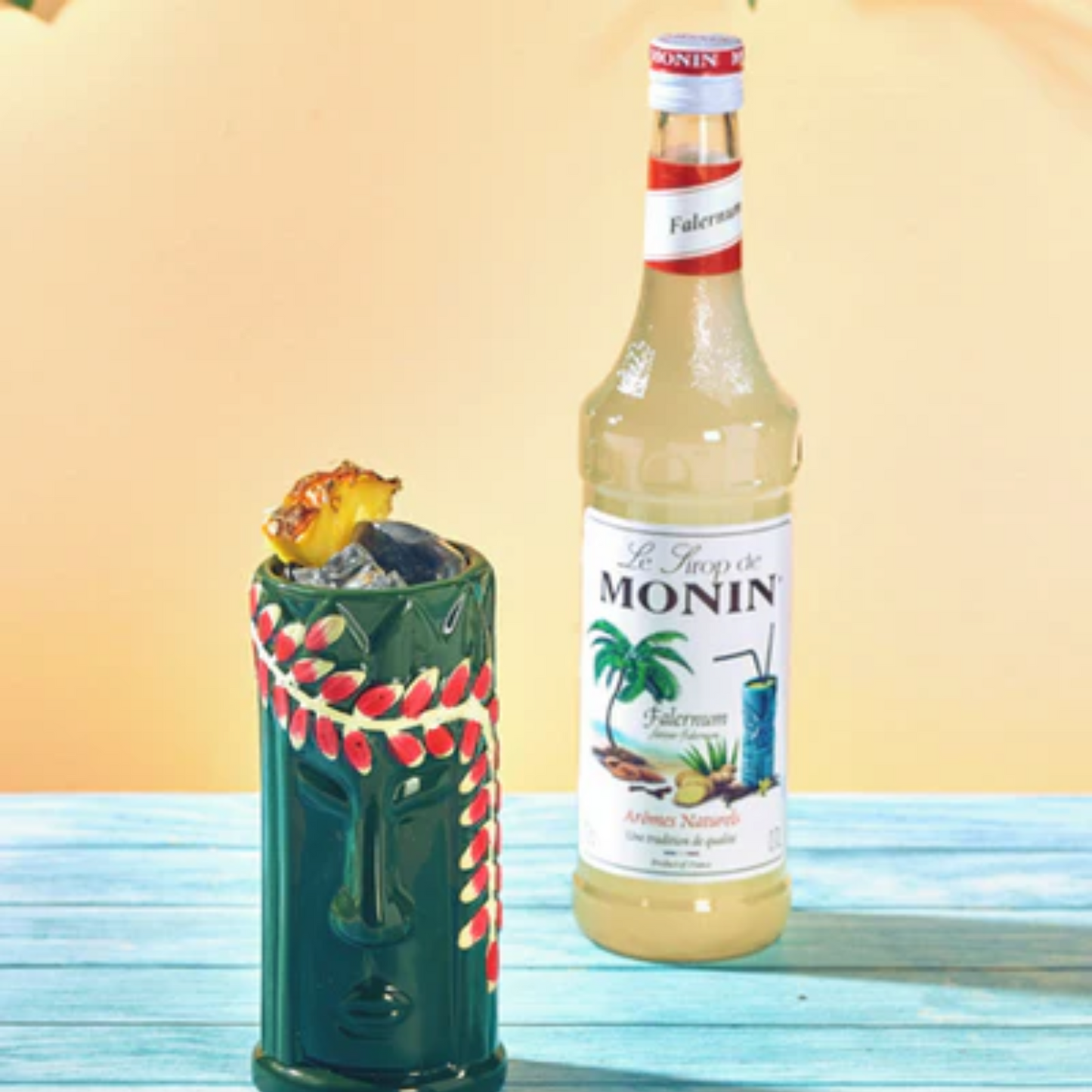 Tropische foto van een Tiki mok op een zomerse blauw houten tafel naast een fles Monin Falernum. Tiki mok is groen met rode decoratie en op de mok ligt een schijfje ananas. Compleet Monin assortiment verkrijgbaar bij Sherlocks Place Rotterdam