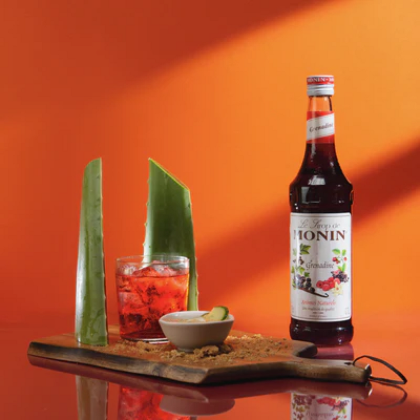 Een zomerse, alcoholvrije cocktail met een helderrode kleur, geserveerd in een glas met ijsblokjes en gepresenteerd op een houten plank met twee verse aloë vera-bladeren. Ernaast staat een fles Monin grenadinesiroop. De cocktail bevat grenadine, citroenconcentraat en ananassap, en wordt geserveerd met een tropische presentatie voor een verfrissende ervaring. Monin verkrijgbaar bij Sherlocks Place Rotterdam