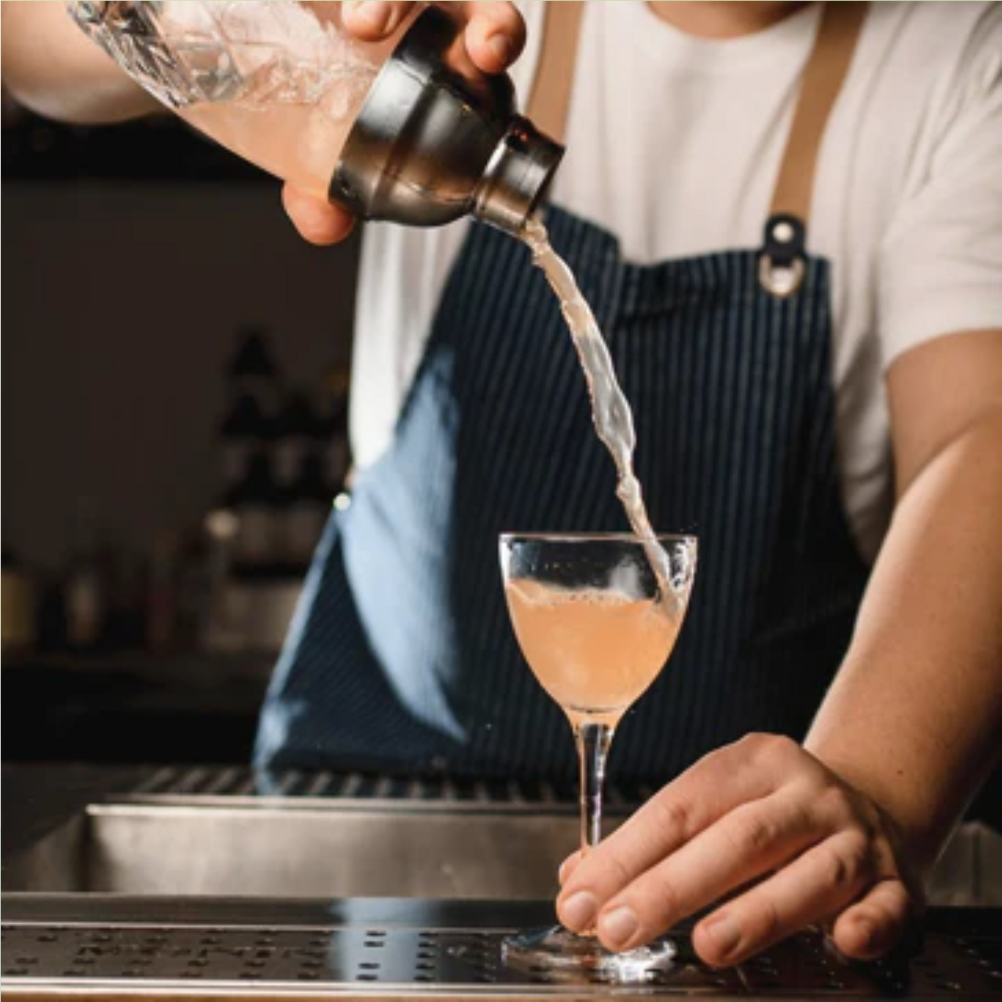 Barman schenkt een frisse Cosmo Lychee met Pamplemousse Rose in een cocktailglas, gemaakt met Monin Lychee en Monin Pamplemousse Rose Siroop – nu verkrijgbaar bij Sherlock’s Place.