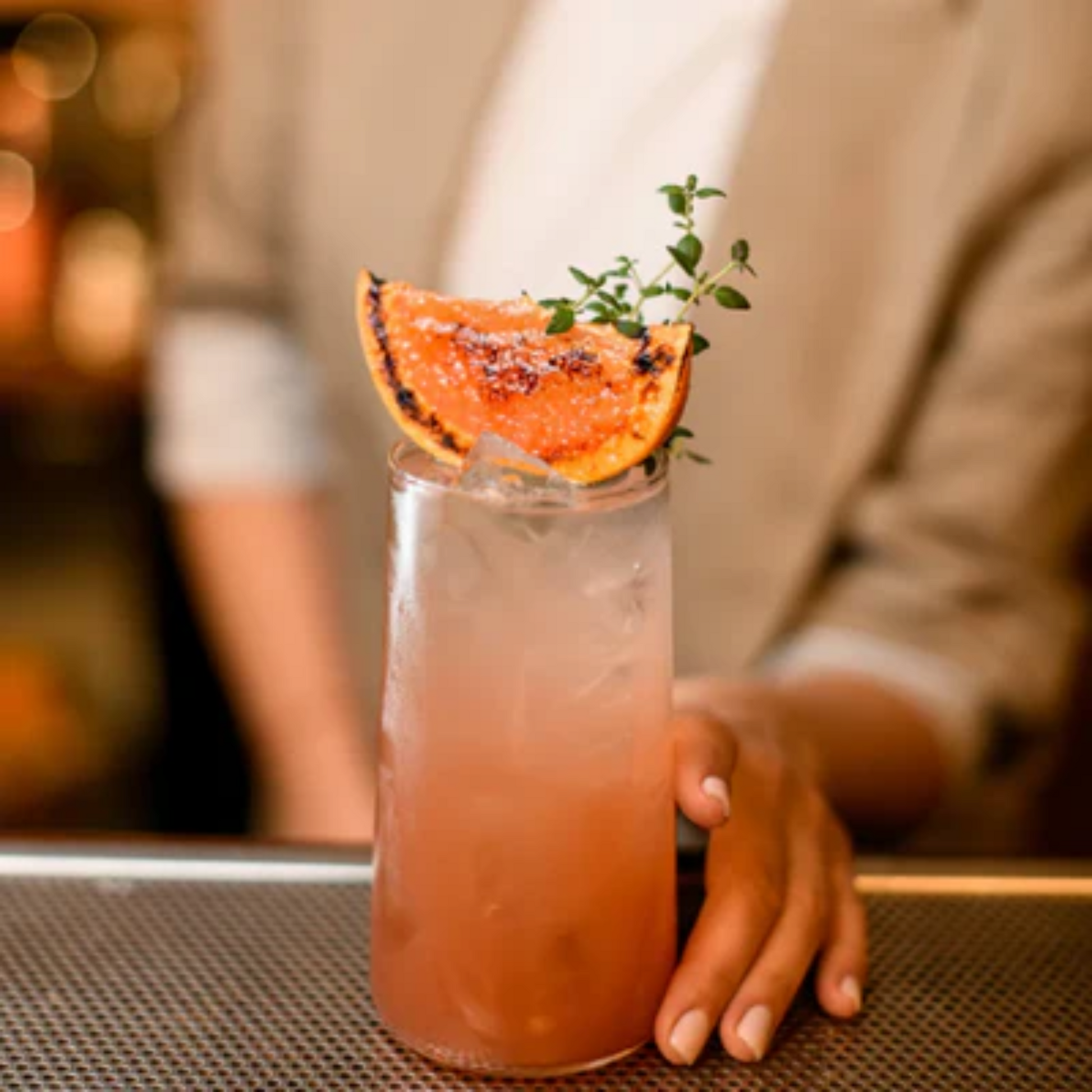 Een Longdrink glas met een Collins Cocktail gemaakt van onder meer Monin Cassis. Op de achtergrond vaag de vrouw die het glas vasthoudt en al garnering op de cocktail een part geroosterde sinaasappel. Monin verkrijgbaar bij Sherlocks Place Rotterdam 