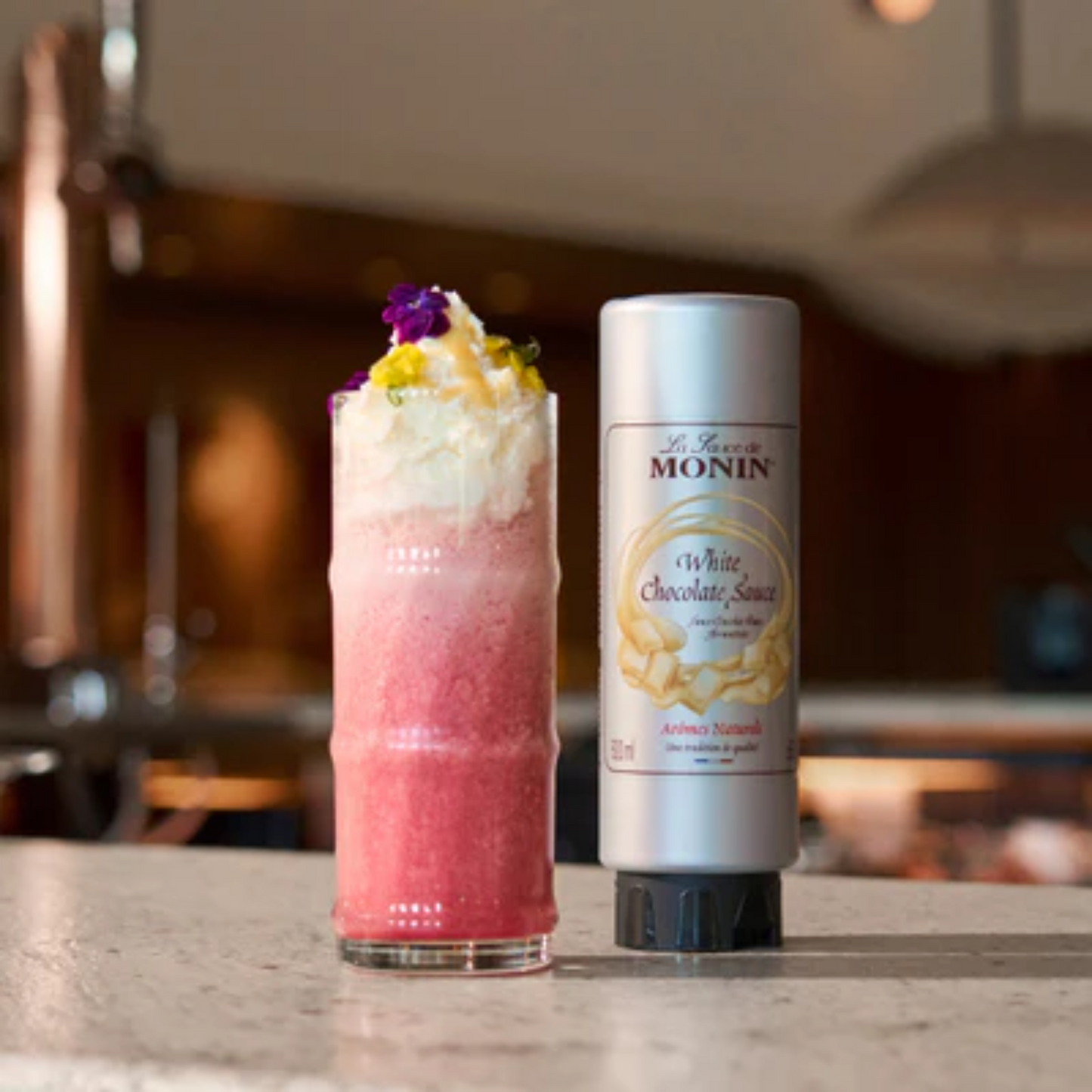 Frisse frambozenmilkshake met slagroom, afgewerkt met eetbare bloemen en Monin White Chocolate Sauce op een marmeren bar, verkrijgbaar bij Sherlock’s Place Rotterdam.