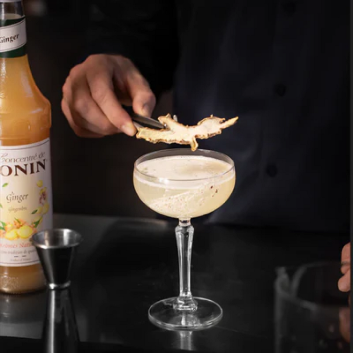 Een bartender garneert een Ginger Margarita met een gedroogd stukje gember, gemaakt met Le Concentré de Monin Ginger. De fles staat op de voorgrond op een zwarte bar, samen met een jigger. Compleet Monin assortiment is verkrijgbaar bij Sherlocks Place in Rotterdam.