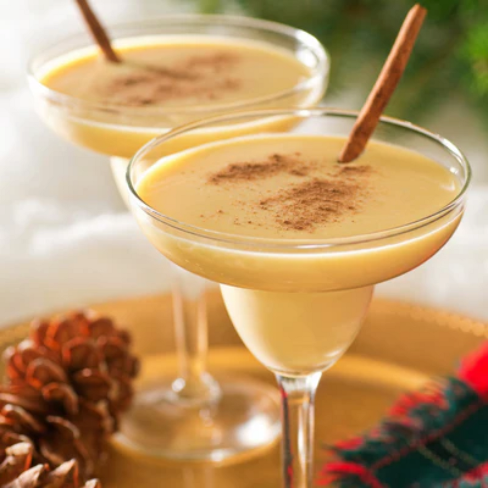 Twee glazen gevuld met romige, gele eggnog, gegarneerd met kaneelpoeder en een kaneelstokje. De glazen staan op een gouden dienblad met dennenappels en een rood-groene plaid. Een perfecte feestelijke sfeer. Compleet Monin assortiment verkrijgbaar bij Sherlocks Place Rotterdam 