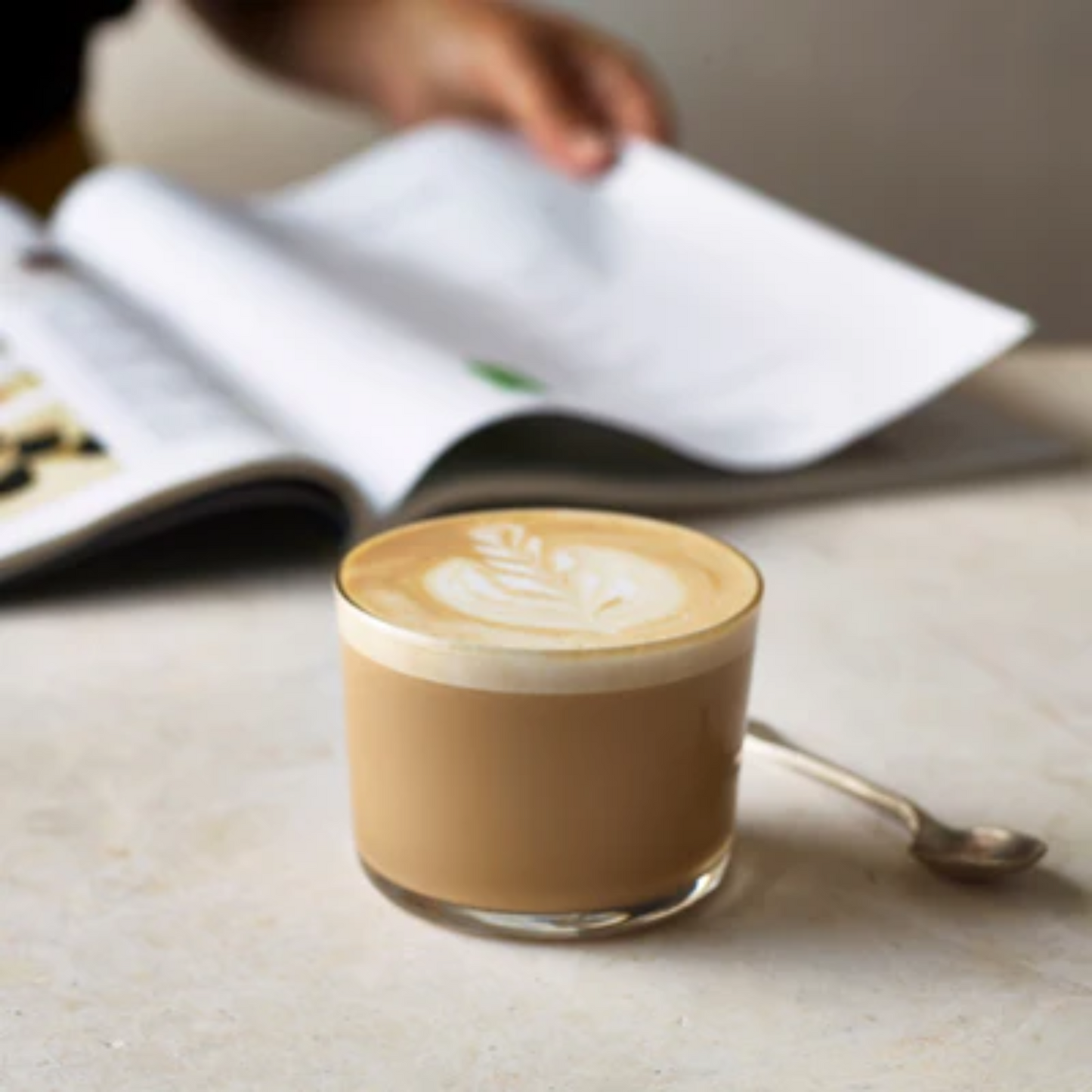 Een romige latte met latte art in een laag glas staat op een lichte marmeren tafel, met een zilveren lepeltje ernaast. Op de achtergrond bladert iemand door een open tijdschrift. Deze latte is op smaak gebracht met Le Sirop de Monin French Vanille en is verkrijgbaar bij Sherlock’s Place Rotterdam.