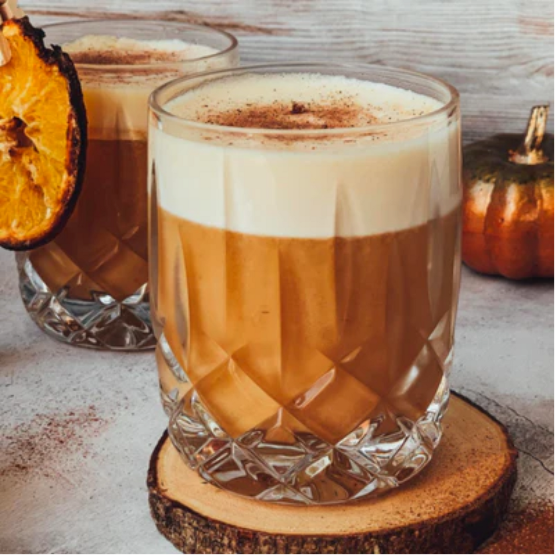 Prachtige gedetaillerde foto van twee glazen met Bourbon Sour Cinnamon Roll  drankjes. Voorste glas staat op een houten ronde onderzetter. De Bourbon Sour heeft een mooie schuimige laag en een schijf geroosterde sinaasappel ter garnering. Monin Verkrijgbaar bij Sherlocks Place Rotterdam