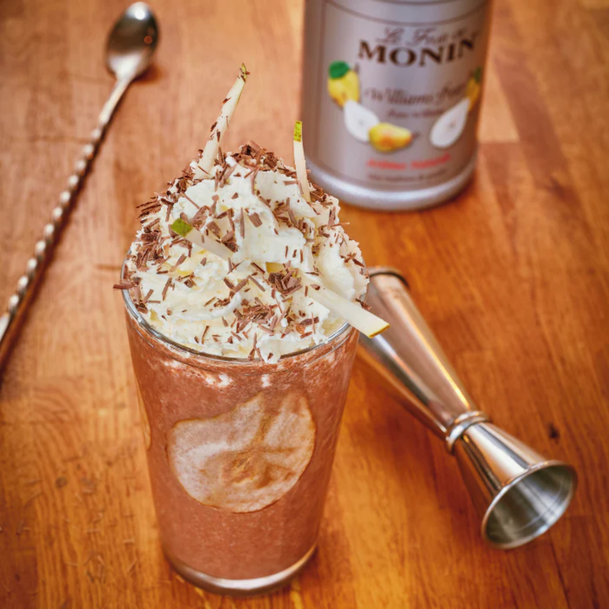 Chocolade frappé met MONIN Chocoladesiroop, afgewerkt met slagroom, chocoladeschaafsel en peer, geserveerd in een glas bij een houten bar – perfecte verwennerij voor koffieliefhebbers en cocktailfans. Verkrijgbaar bij Sherlocks Place otterdam