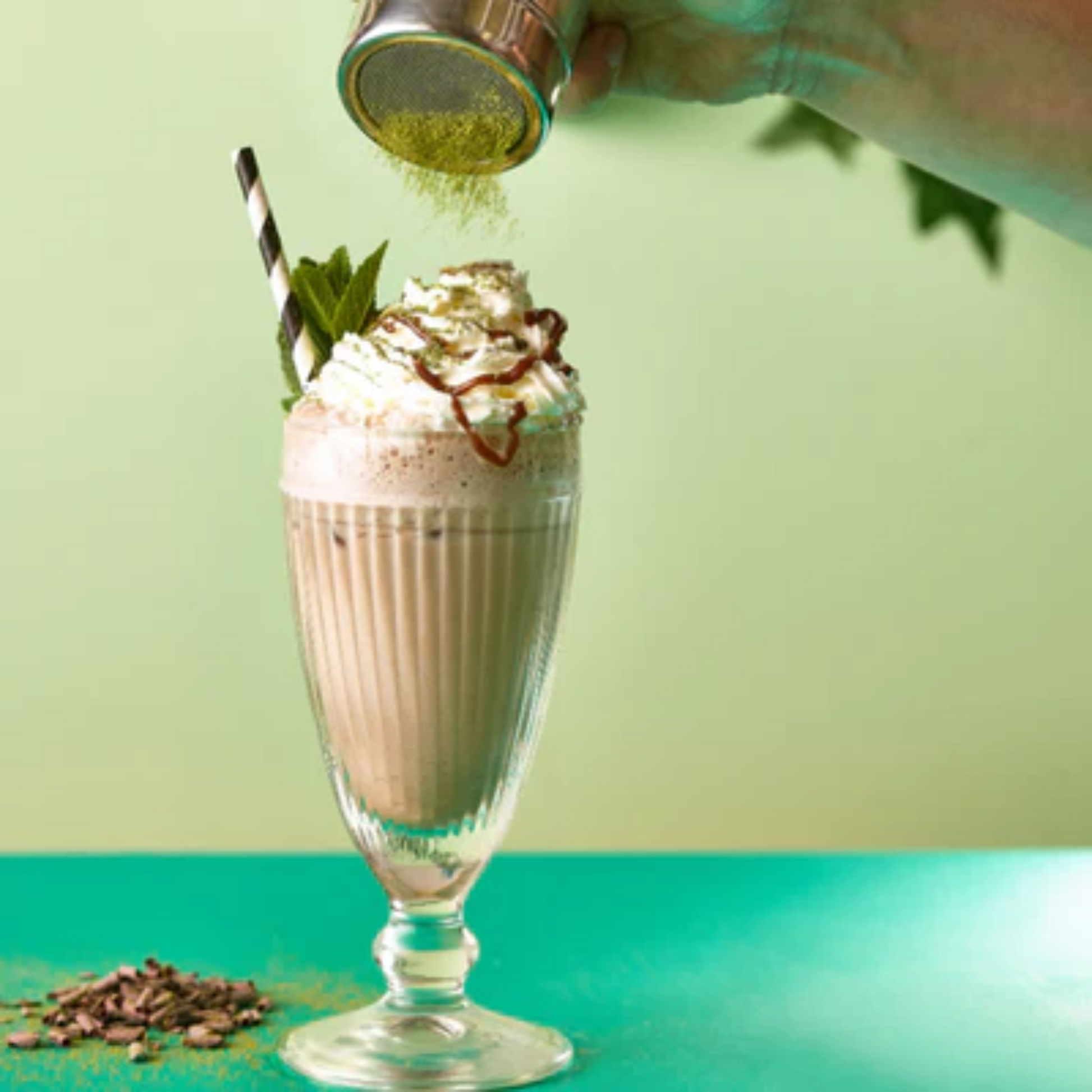 Chocolade Pistache frappé met MONIN Chocoladesiroop, Pistache siroop en Vanille Frappe afgewerkt met slagroom, chocoladeschaafsel en munt en cacaopoeder, geserveerd in een glas bij een houten bar – perfecte verwennerij voor koffieliefhebbers en cocktailfans.