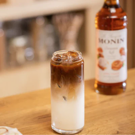 Stijlvolle presentatie van een fles Monin Cinnamon Roll 70cl naast een Cold Brew Latte met Cinnamon Roll en Caramel op een houten tafel. Minimalistische opstelling. Musthave voor koffieliefhebbers! Verkijgbaar bij Sherlocks Place Rotterdam