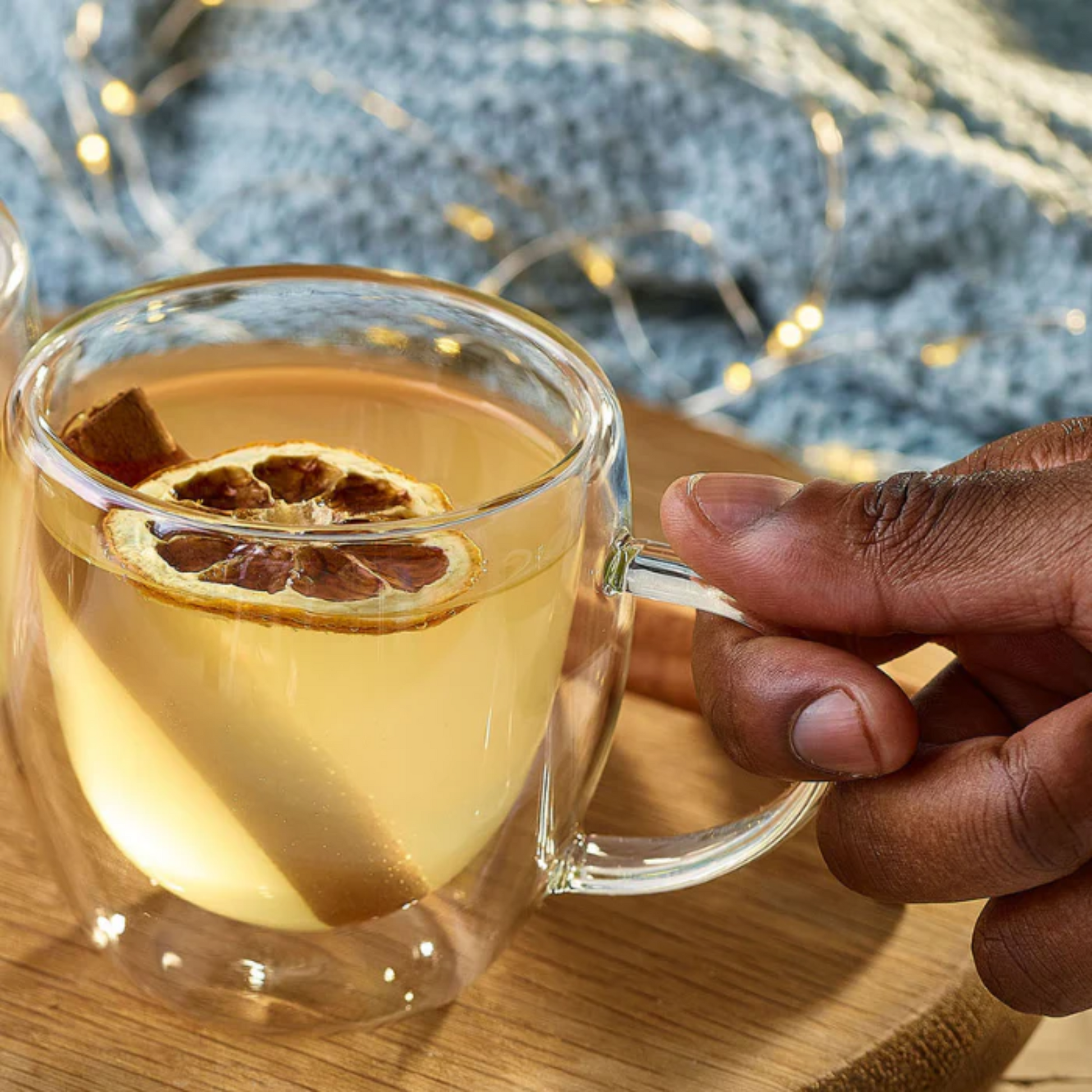 Een mooie close-up foto van een transparant theeglas wat wordt neergezet op een tafel. Een deel van de hand is zichtbaar. De Thee is gemaakt met Monin Lime en Passion Fruit. In het glas zit ook nog een kaneelstokje en een schijfje gedroogde citroen. Perfect drankje voor koude maanden om je immuunsysteem een oppepper te geven! Verkrijgbaar bij Sherlocks Place Rotterdam