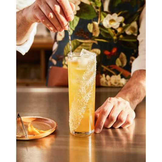 Close-up foto van een White Tea Mango Cocktail gemaakt met Monin. Het is een chique foto met op de achtergrond de bartender met een fleurig schort die de cocktail aan het bereiden is. Hij heeft een bar spoon in de hand. Op de voorgrond het glas; een longdrink gevuld met ijsklontjes en de Monin ingrediënten. De kleur is oranje. Ernaast een schaaltje met daarop een pincet en slices citrus. Alles van Monin verkrijgbaar bij Sherlocks Place Rotterdam