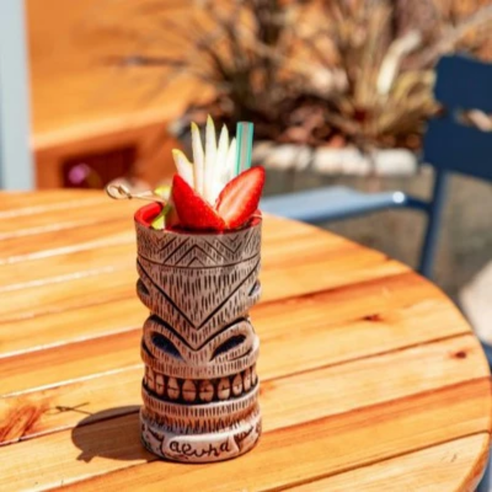 Tropische Tiki mok, bruin, gepositioneerd op een ronde houten tafel. Veel zonlicht Afgetopt met aardbeien. Monin Strawberry perfect voor Tiki's, Lemonades, ijscreaties, en zo veel meer. Verkrijgbaar bij Sherlocks Place Rotterdam