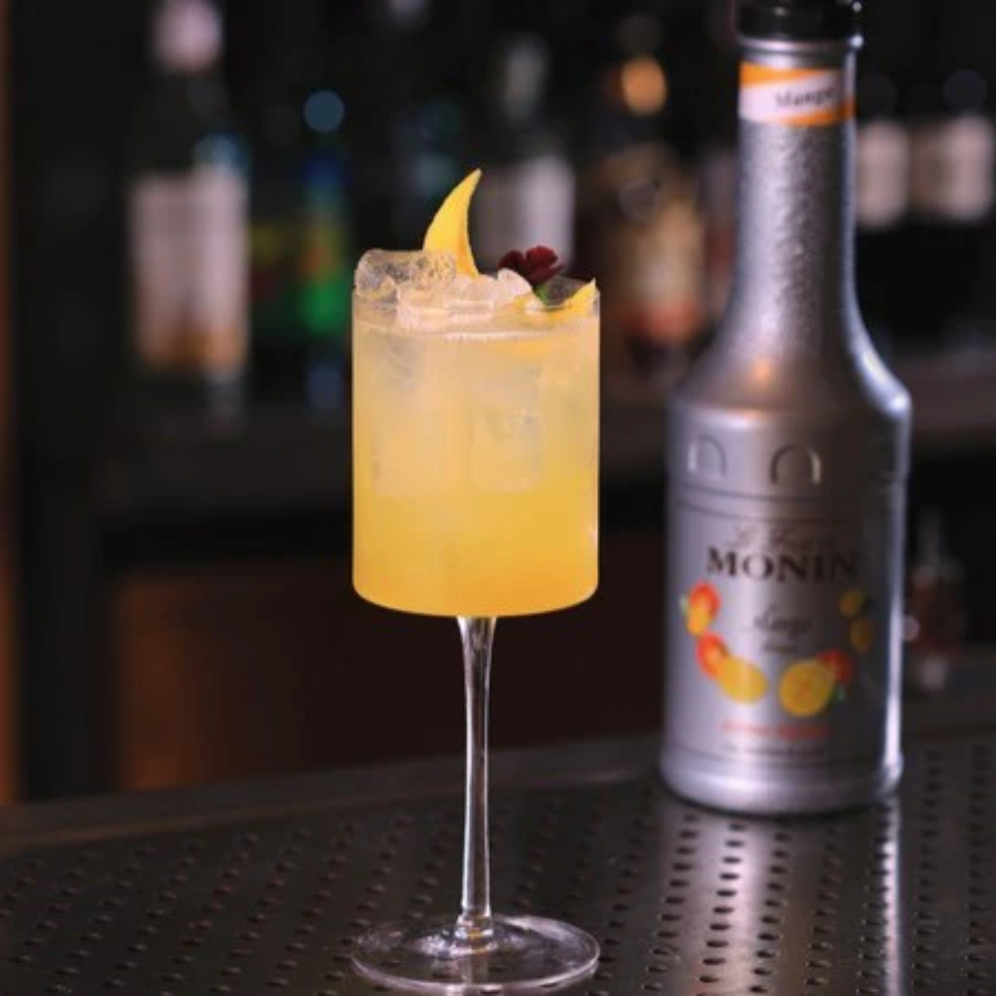 Mango Elderflower Lemonade geserveerd in een strak wijnglas en gegarneerd met citrus en eetbare bloemen. Geplaatst op een bar met op de achtergrond een fles Monin Mango  Le Fruit puree. Minimalistische setting. Ideaal voor Virgin Cocktails en alcoholvrije drankjes liefhebbers. Compleet Monin Assortiment verkrijgbaar bij Sherlocks Place Rotterdam  